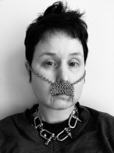 Black and white selfie photo of Theo, a white person with dark hair, wearing a chain maille moustache